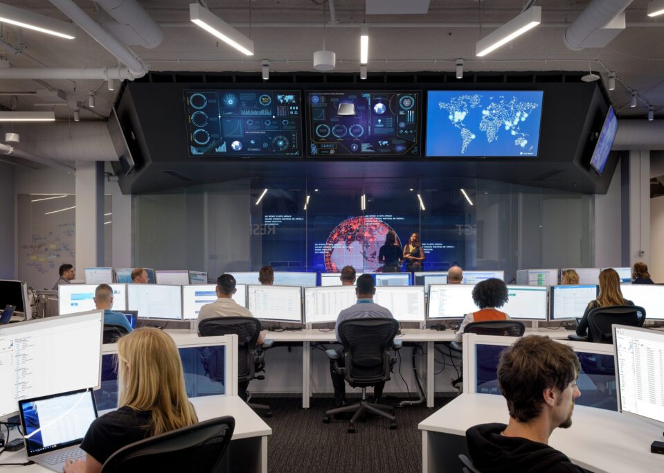 Work room with people at desks and screens in the background
