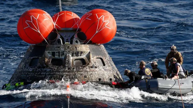 The Orion capsule floats in the Pacific Ocean while Navy personnel secure the cable from the raft.