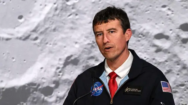 A middle-aged man wearing a black NASA jacket and red tie speaks while looking slightly off camera, with a huge image of the Moon's surface in the background.