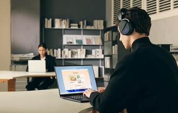 A man in profile with black headphones works on a dark laptop, hands on the keyboard. In the background, a woman is using a white laptop. There are shelves with books